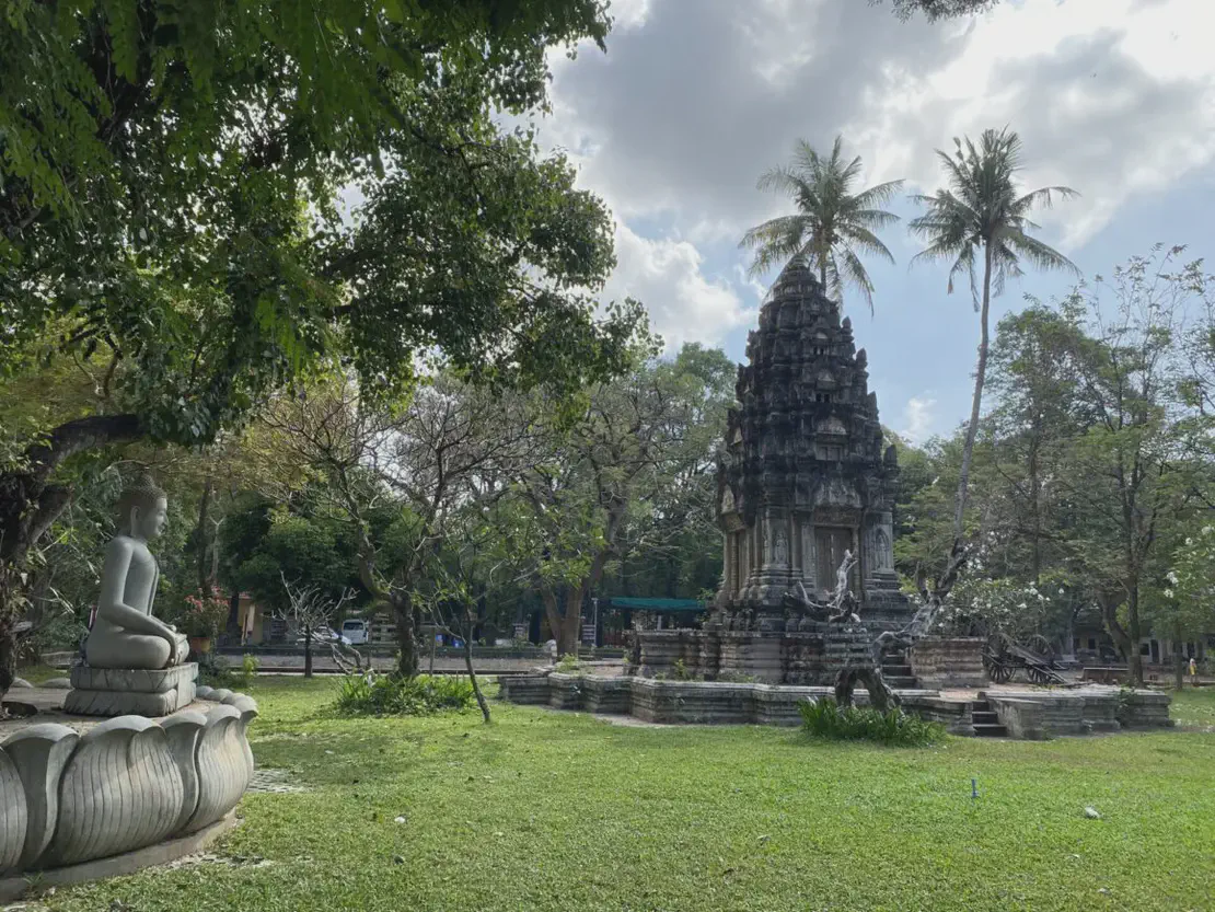 Stone Buddha statue facing an ancient temple structure in a green park with palm trees.
