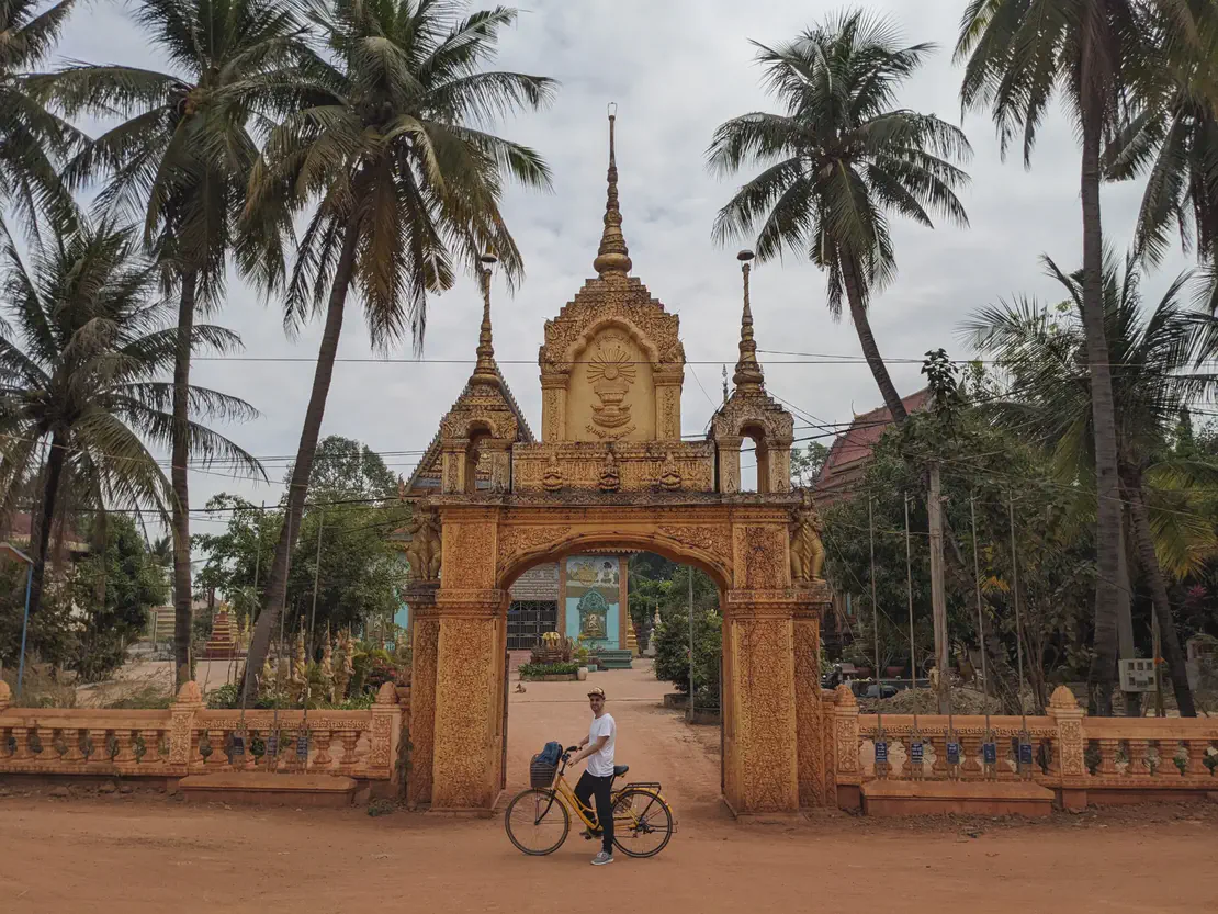 Person with a bicycle standing in front of an ornate golden temple gate adorned with carvings and spires, framed by tall palm trees on a dirt road.