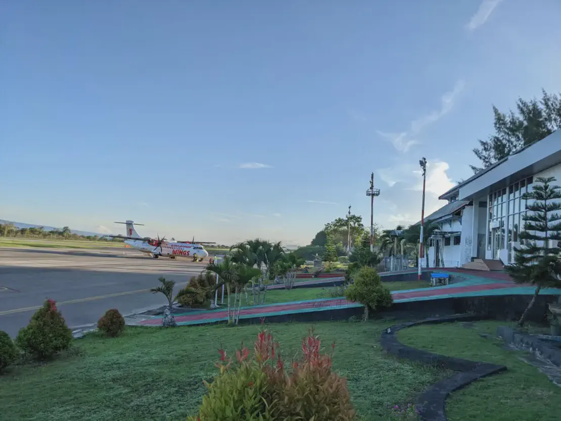 Propeller airplane parked on the runway near a small airport terminal with landscaped greenery in the foreground.