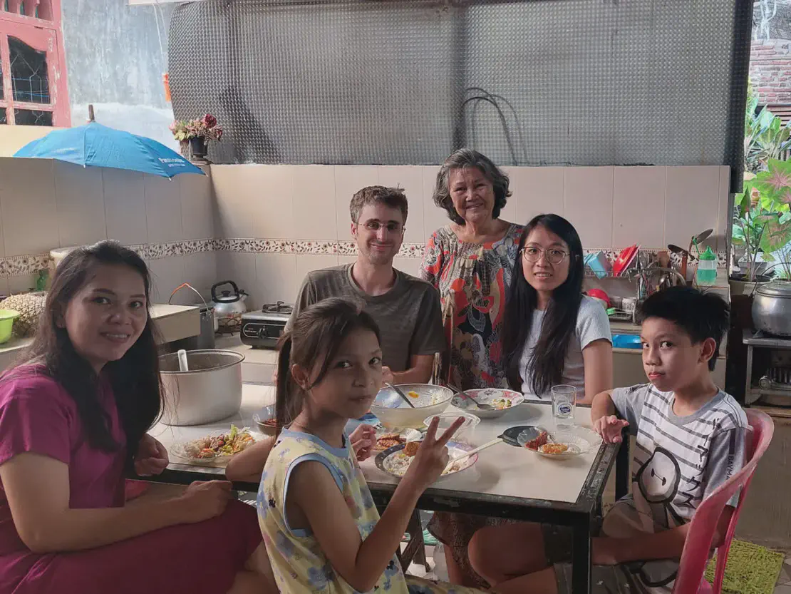 Family meal in a kitchen with six people sitting and smiling at the camera, plates of food on the table and cooking utensils in the background.