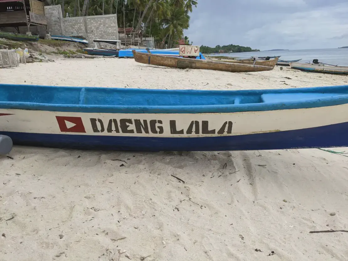 A blue and white wooden boat on the sandy beach with the name 'DAENG LALA' painted on its side.