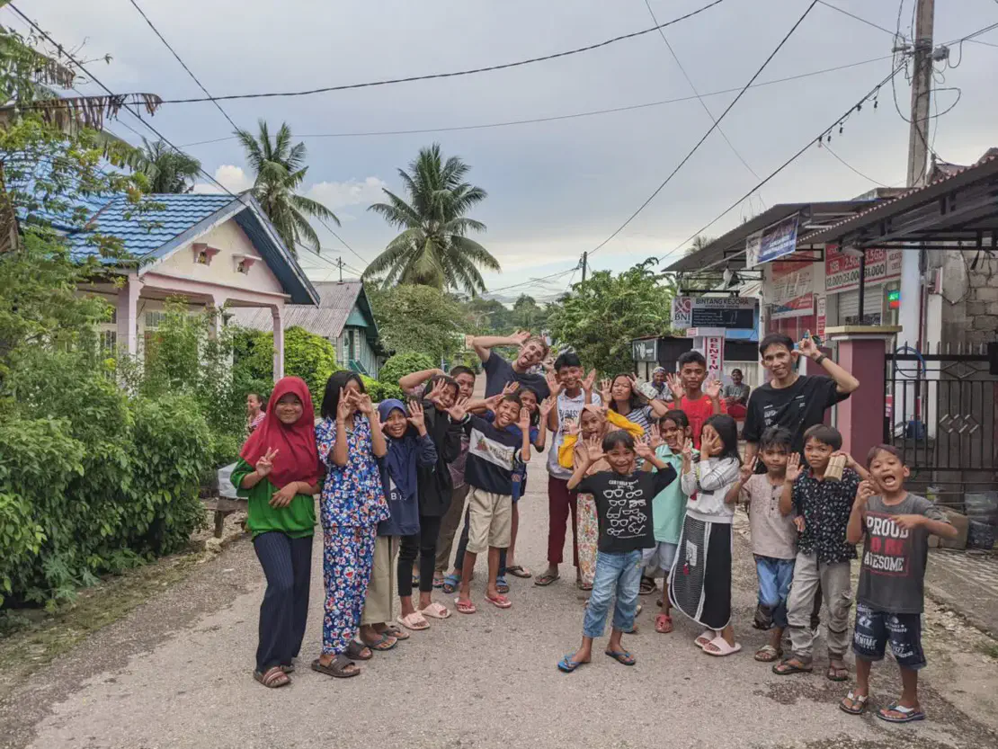 Group of children and a few adults smiling and posing playfully on a village street with houses and palm trees in the background.