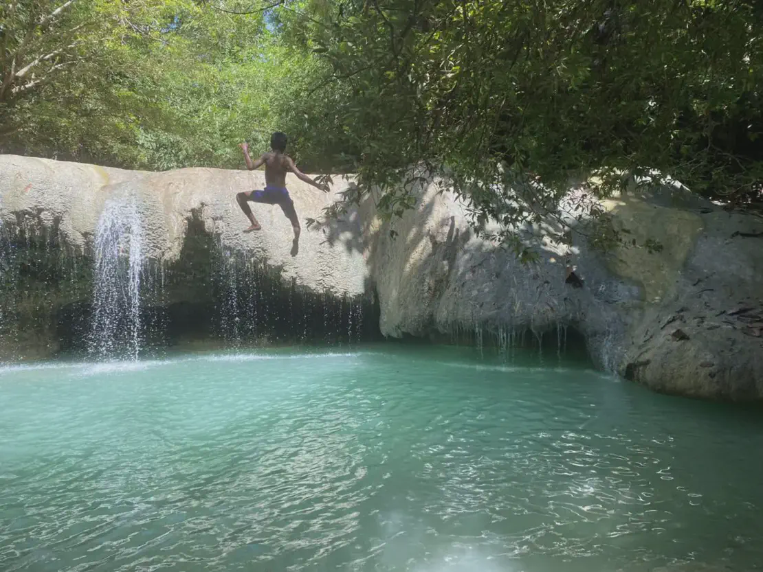 A boy jumping off a rock ledge into a turquoise pool beside a small waterfall, surrounded by lush greenery.