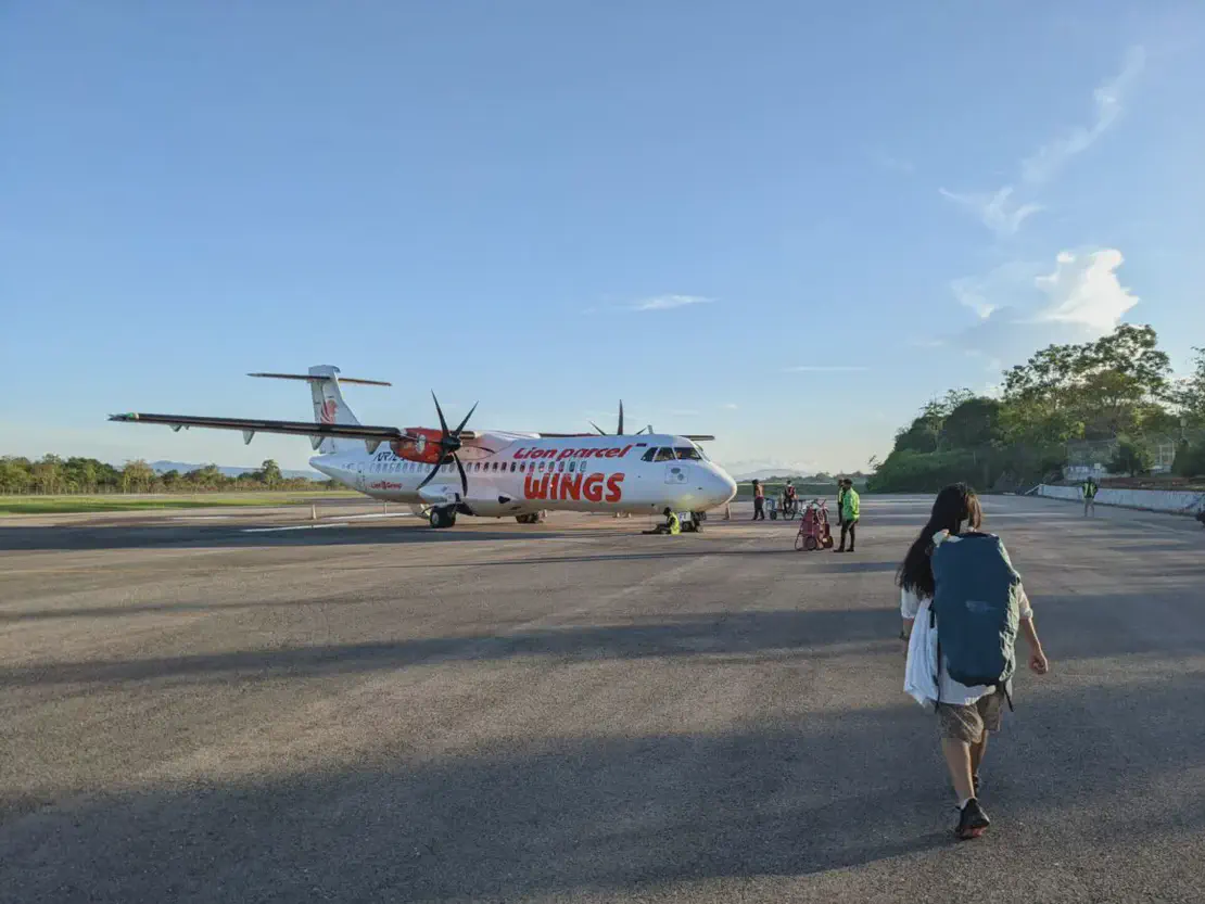 Traveler walking on the airport tarmac with a large backpack toward a Wings Air propeller plane under a clear sky.