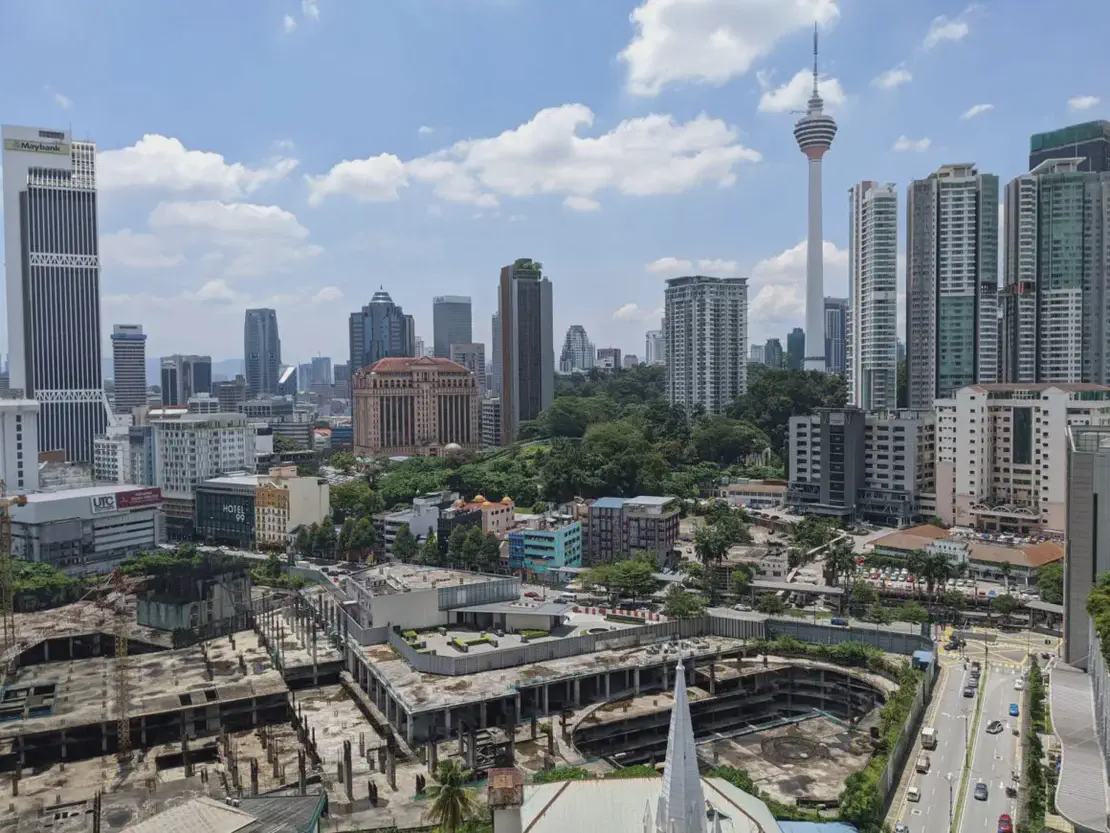 Cityscape of Kuala Lumpur with modern skyscrapers, including the Kuala Lumpur Tower, surrounded by green trees and construction sites in the foreground.
