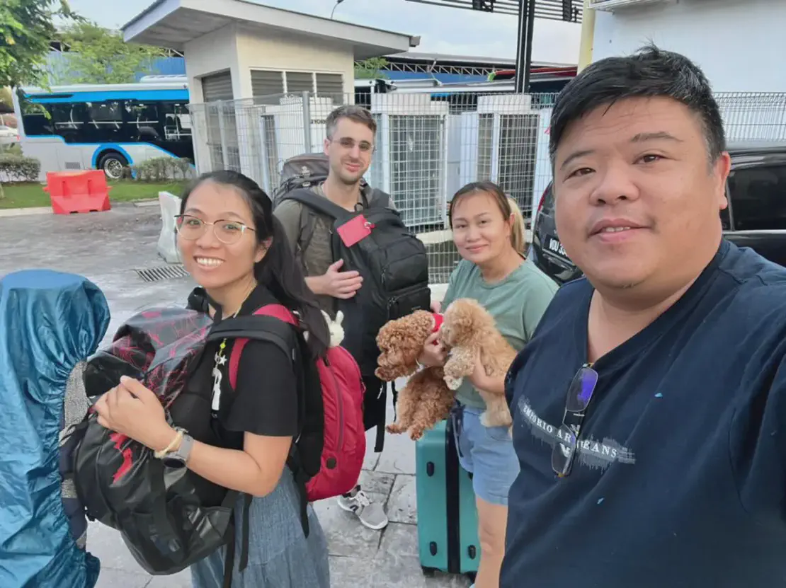Group of four people with luggage and two small dogs outdoors near a bus stop.