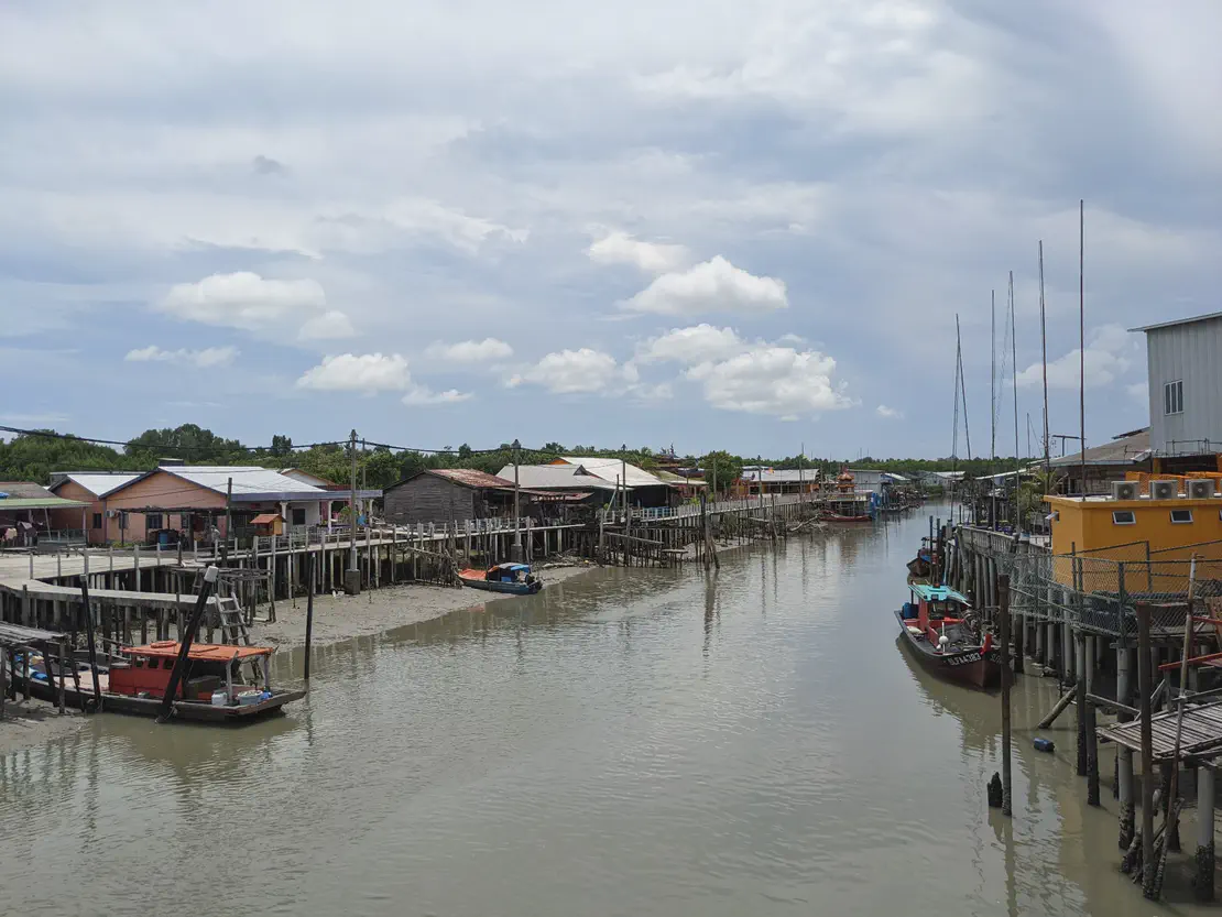 A riverside village with wooden stilt houses and boats moored along the banks under a partly cloudy sky.
