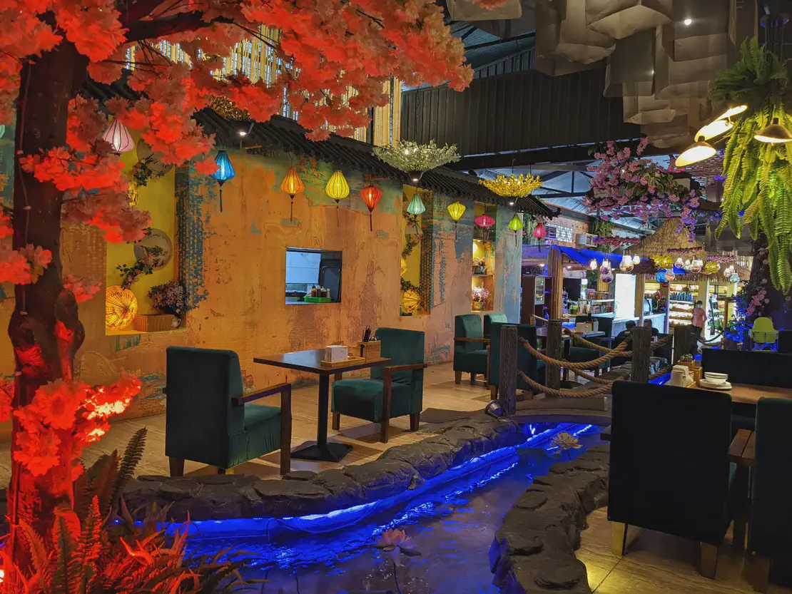 Interior of a themed restaurant with colorful hanging lanterns, artificial cherry blossom trees, and a glowing blue water feature running between dining tables.