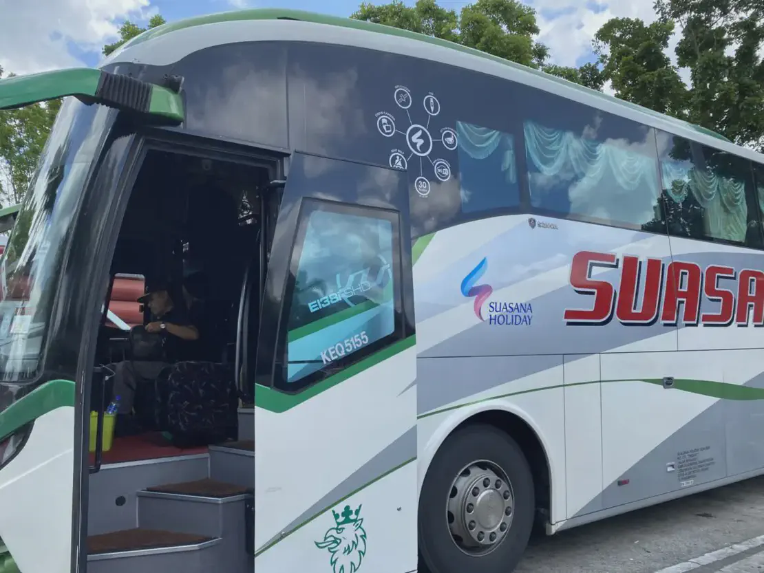 A Suasana Holiday tour bus parked with its door open, showing the driver seated inside. The bus has a white and green exterior with large tinted windows.