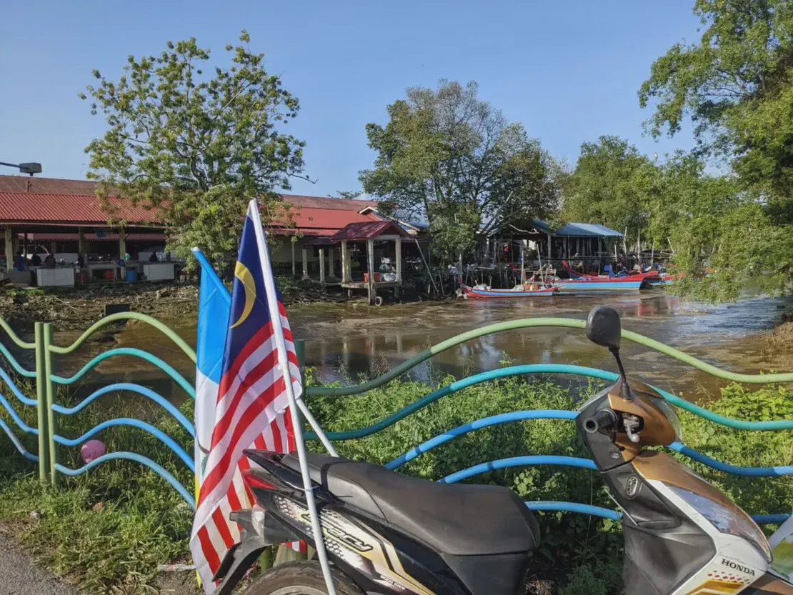 Malaysian flag and Penang flag attached to a motorbike parked by a colorful fence, with a river, fishing boats, and houses on stilts in the background.