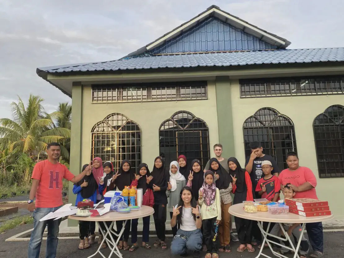 Group of children and adults posing outside a building with tables of food and drinks.