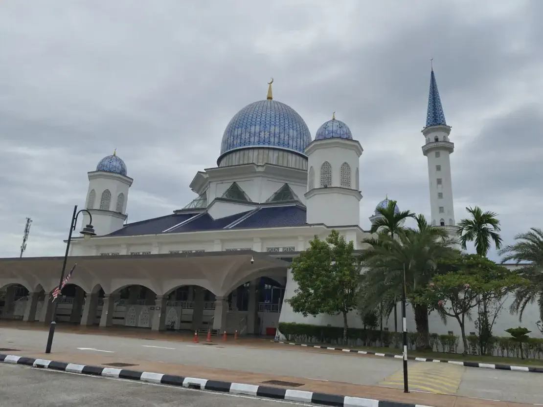 Exterior view of a mosque with blue domes and a tall minaret, surrounded by palm trees under a cloudy sky.