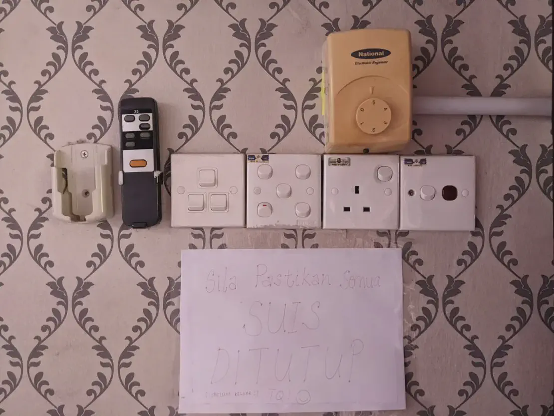 Wall with patterned wallpaper featuring multiple electrical switches, a National electronic regulator, remote control, and a handwritten sign in Malay reminding to switch off power before leaving.