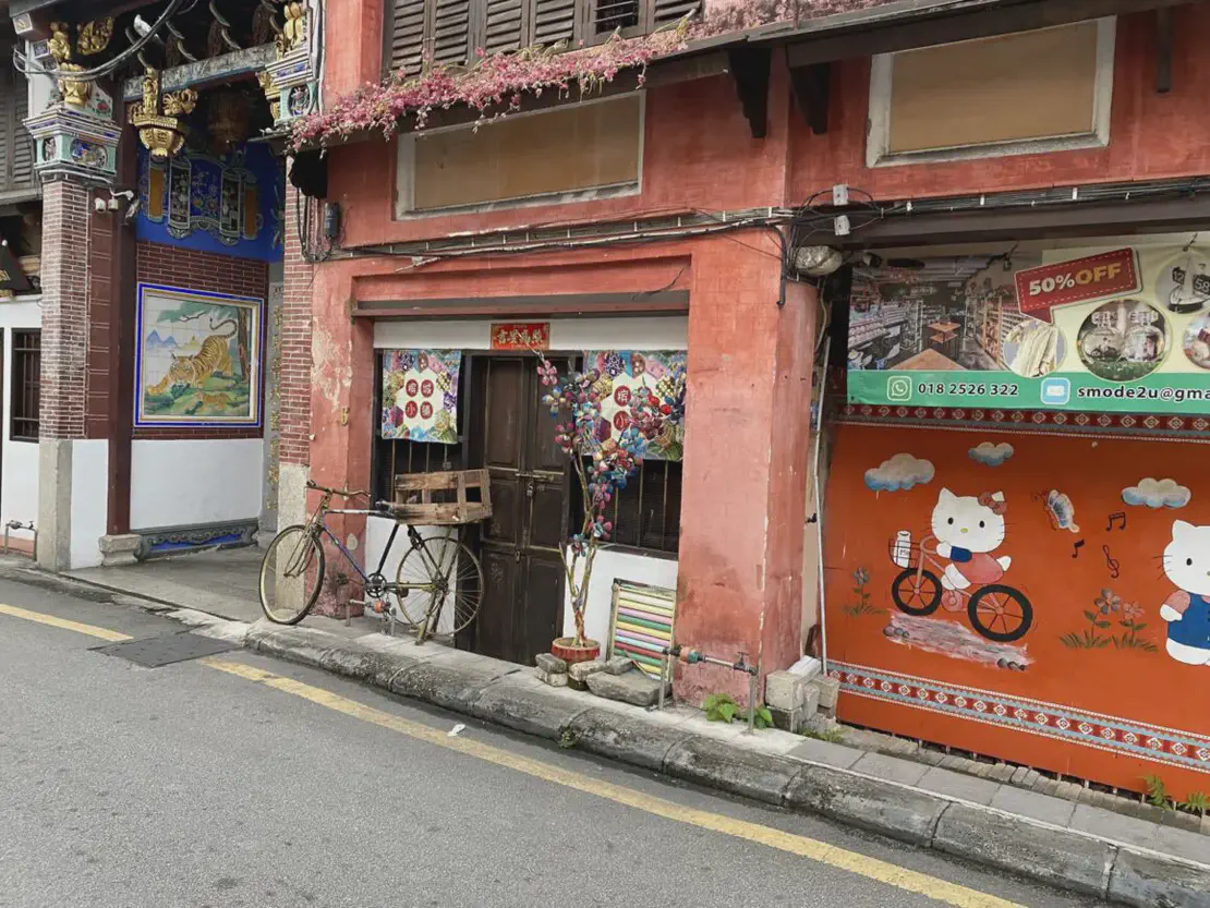 Street view of colorful old buildings with murals, including a tiger painting and Hello Kitty artwork, a bicycle leaning against a wall, and a shopfront decorated with flowers and lanterns.