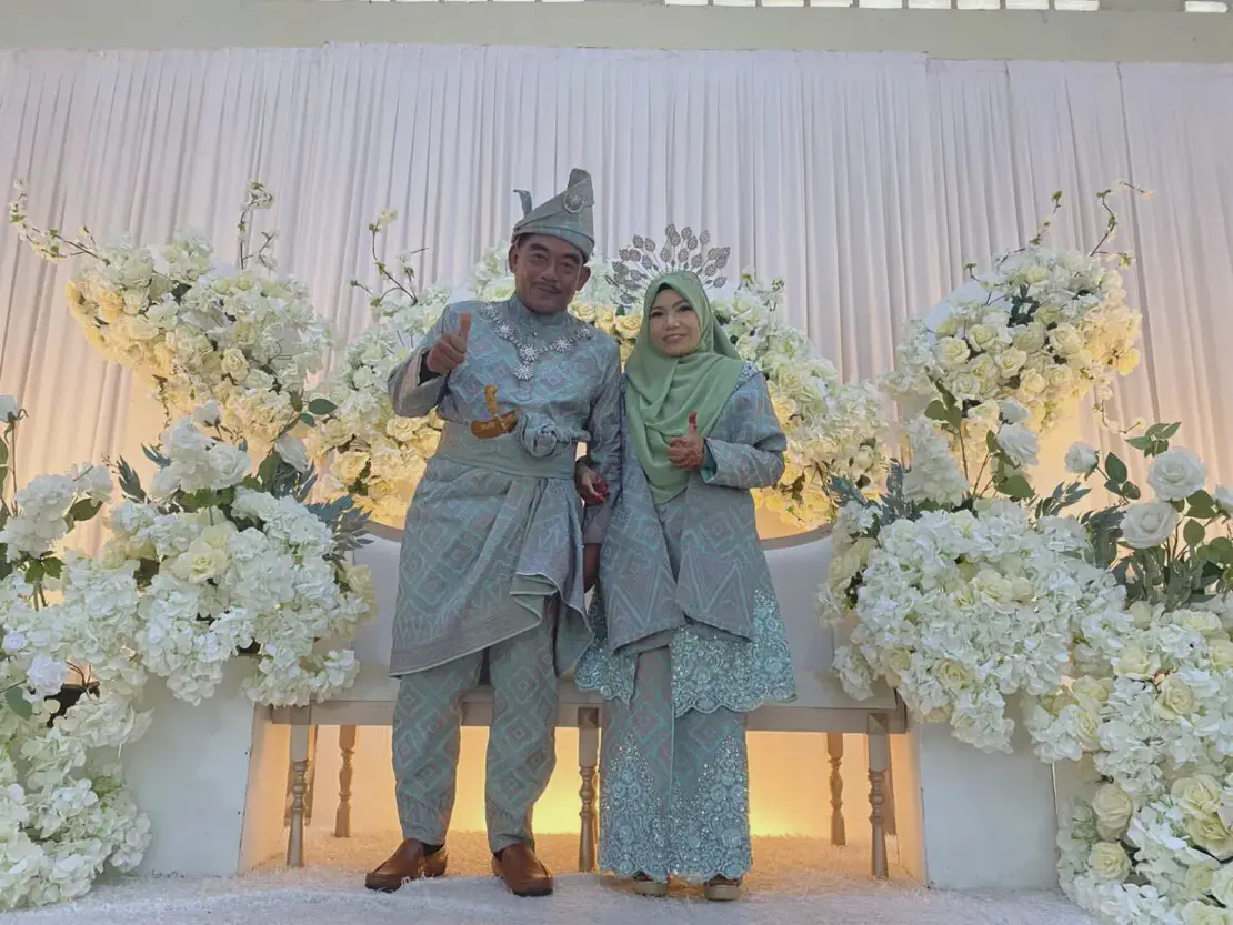 Bride and groom in traditional Malay wedding attire posing on a decorated stage with white flowers.