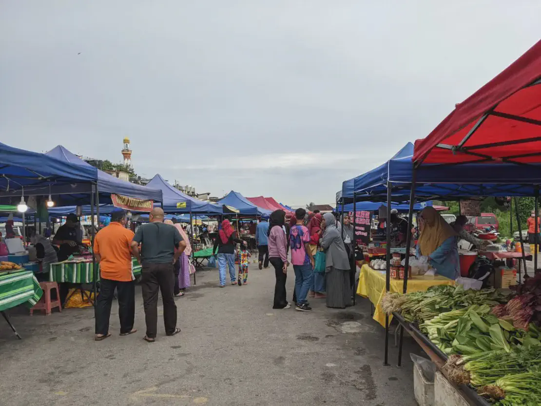 Outdoor street market with rows of colorful canopy stalls selling vegetables and food, vendors interacting with customers, and people walking through the lively marketplace.