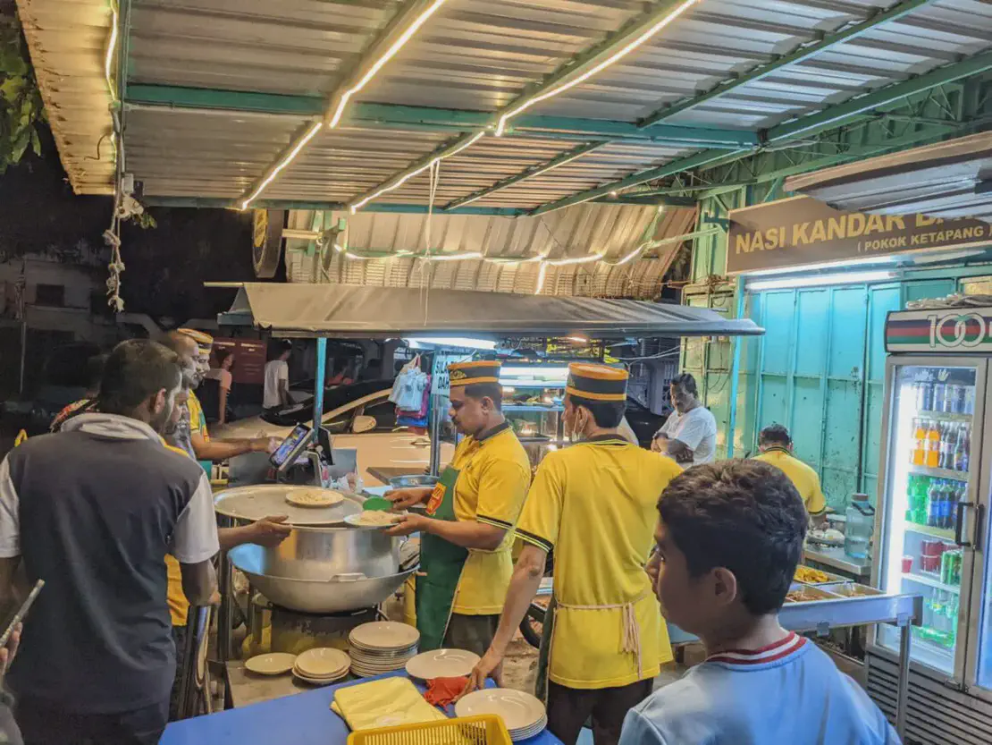 Busy nasi kandar food stall at night with staff in yellow uniforms serving rice and dishes from large pots, while customers wait in line under a lit canopy.