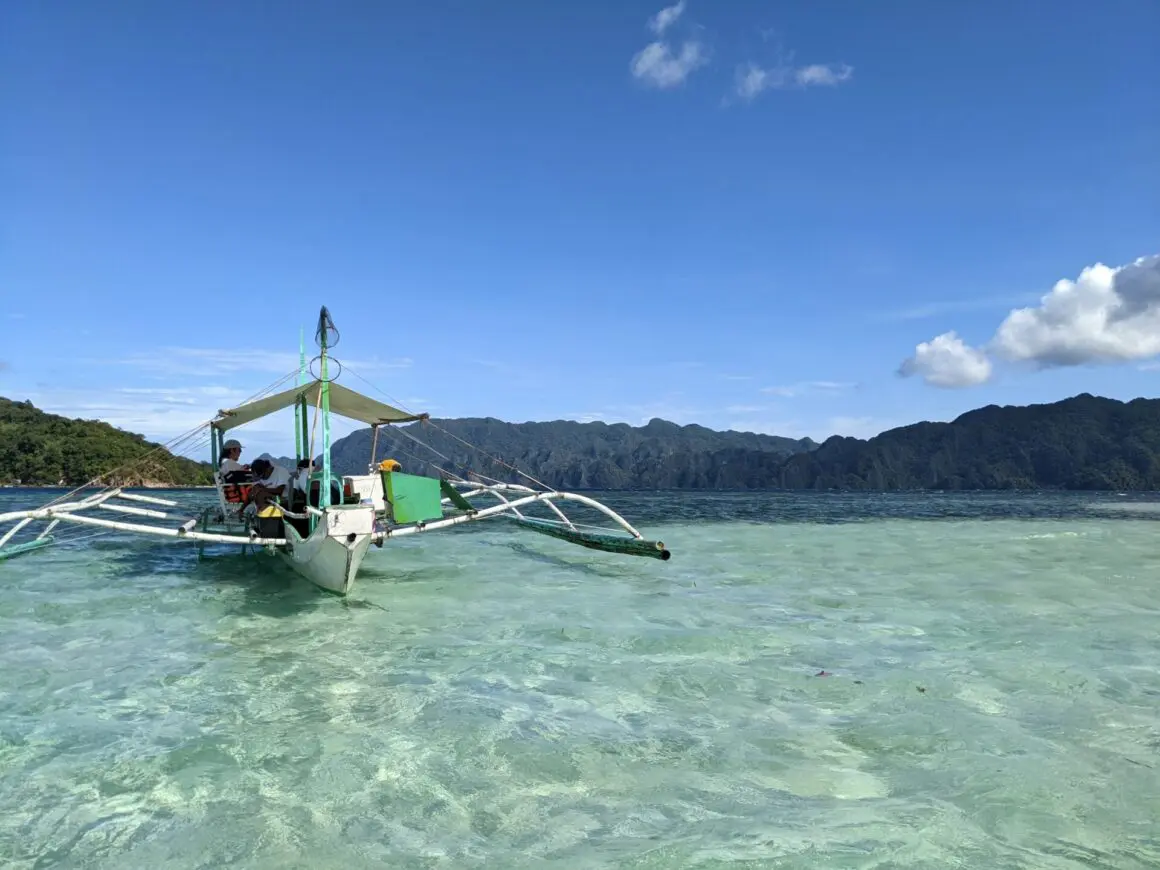 Small outrigger boat floating on clear turquoise water with green mountains in the background under a bright blue sky.