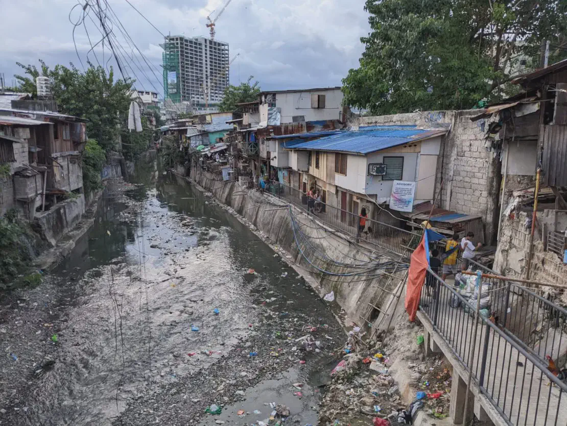 Polluted canal lined with informal houses and scattered trash in an urban area.