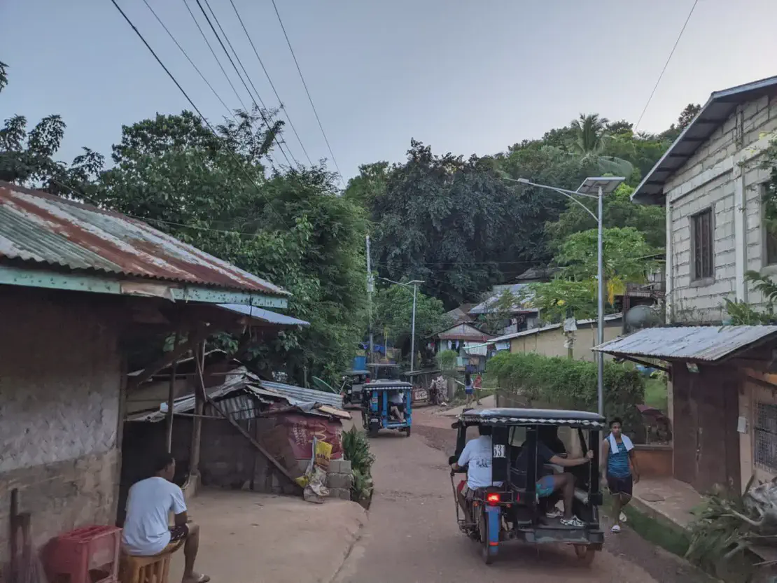 Narrow village street with tricycles, small houses, and people walking in the late afternoon.