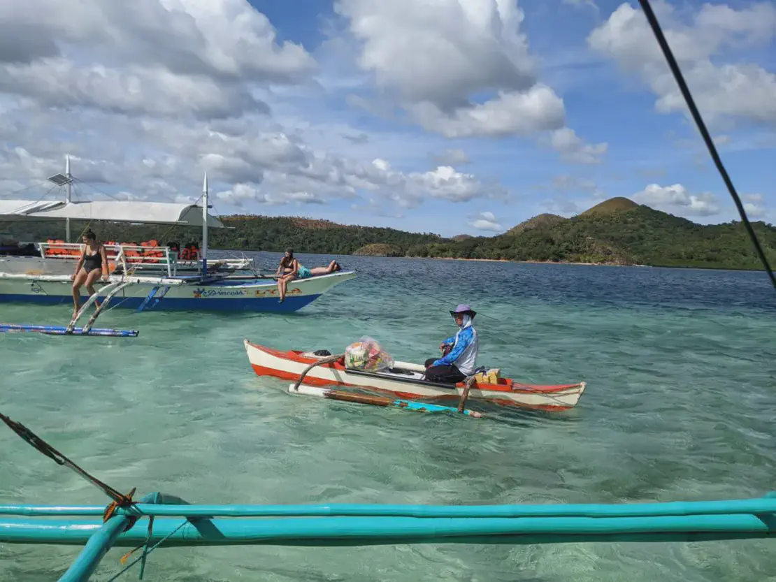Person in a small wooden boat selling goods near a larger tour boat with people relaxing in clear tropical waters.