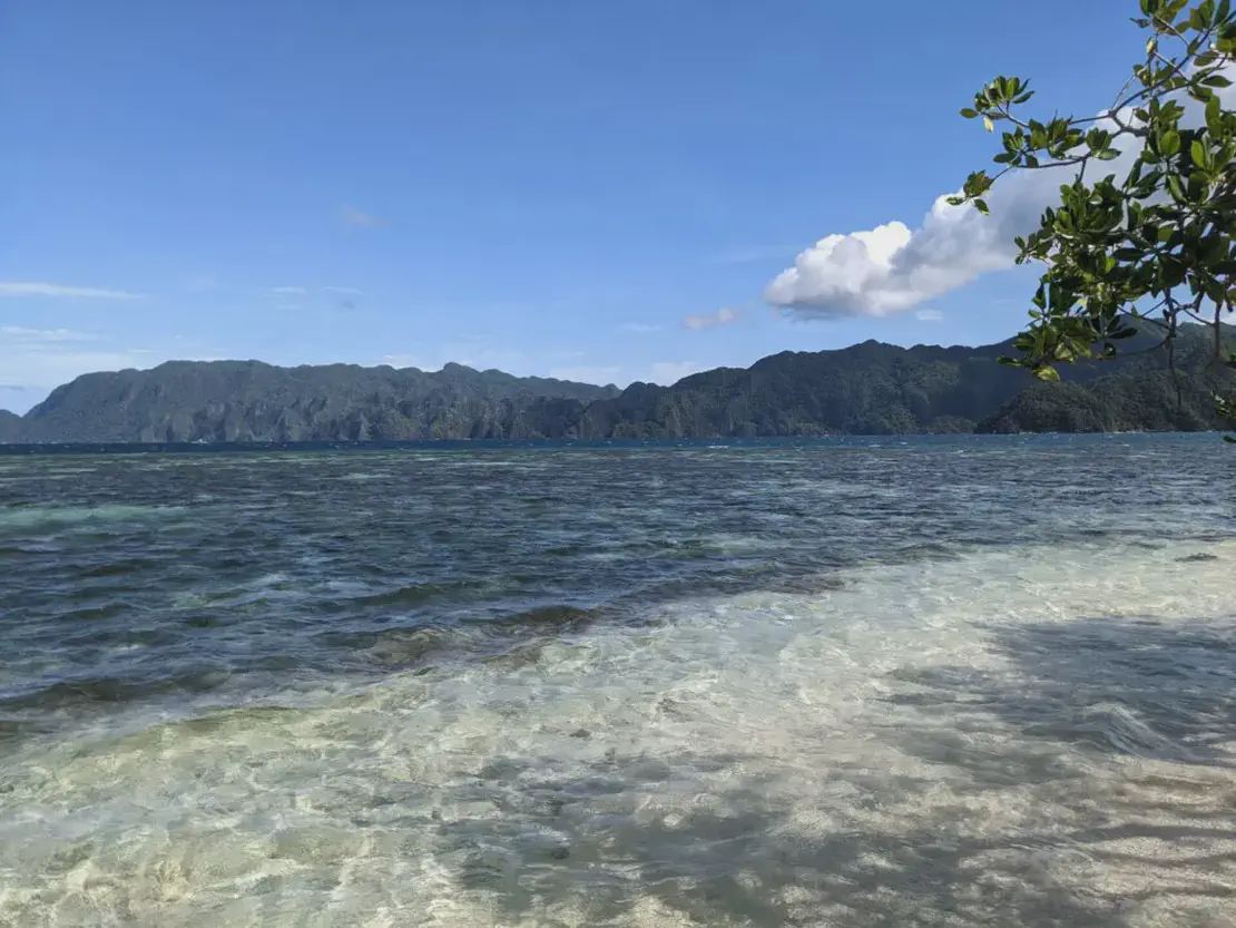 Clear shallow water leading to deep blue sea with forested mountains in the distance under a bright sky.