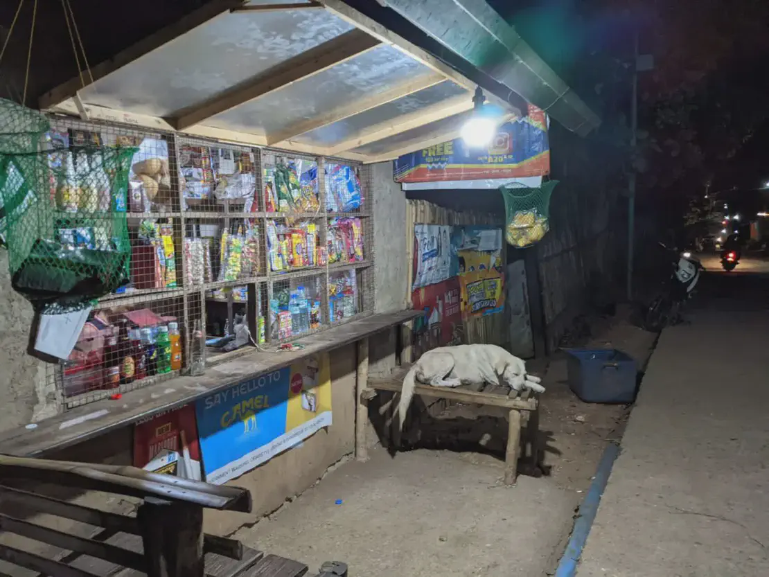 Small roadside store at night with snacks displayed and a dog lying on a wooden bench.