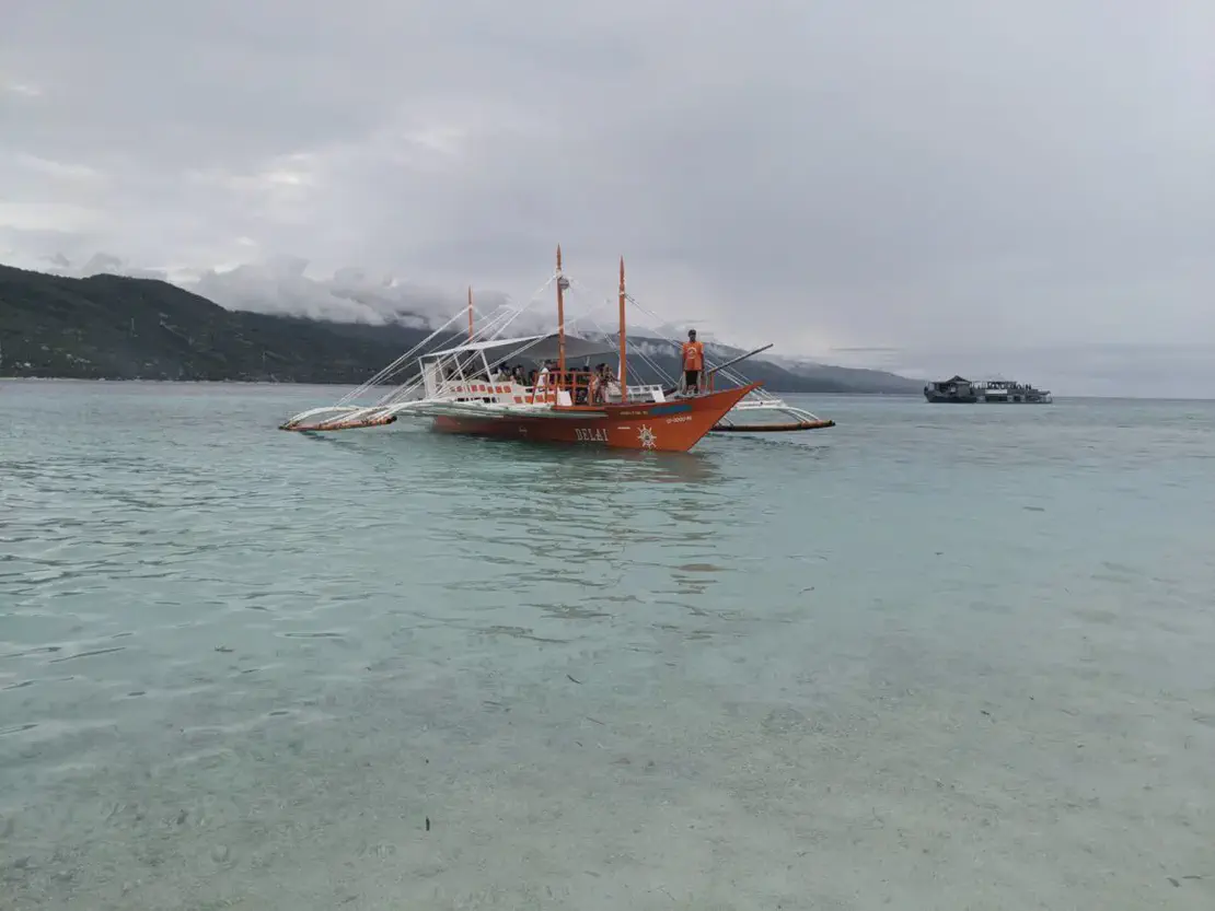 Orange outrigger boat with passengers floating on clear shallow water near the shore.