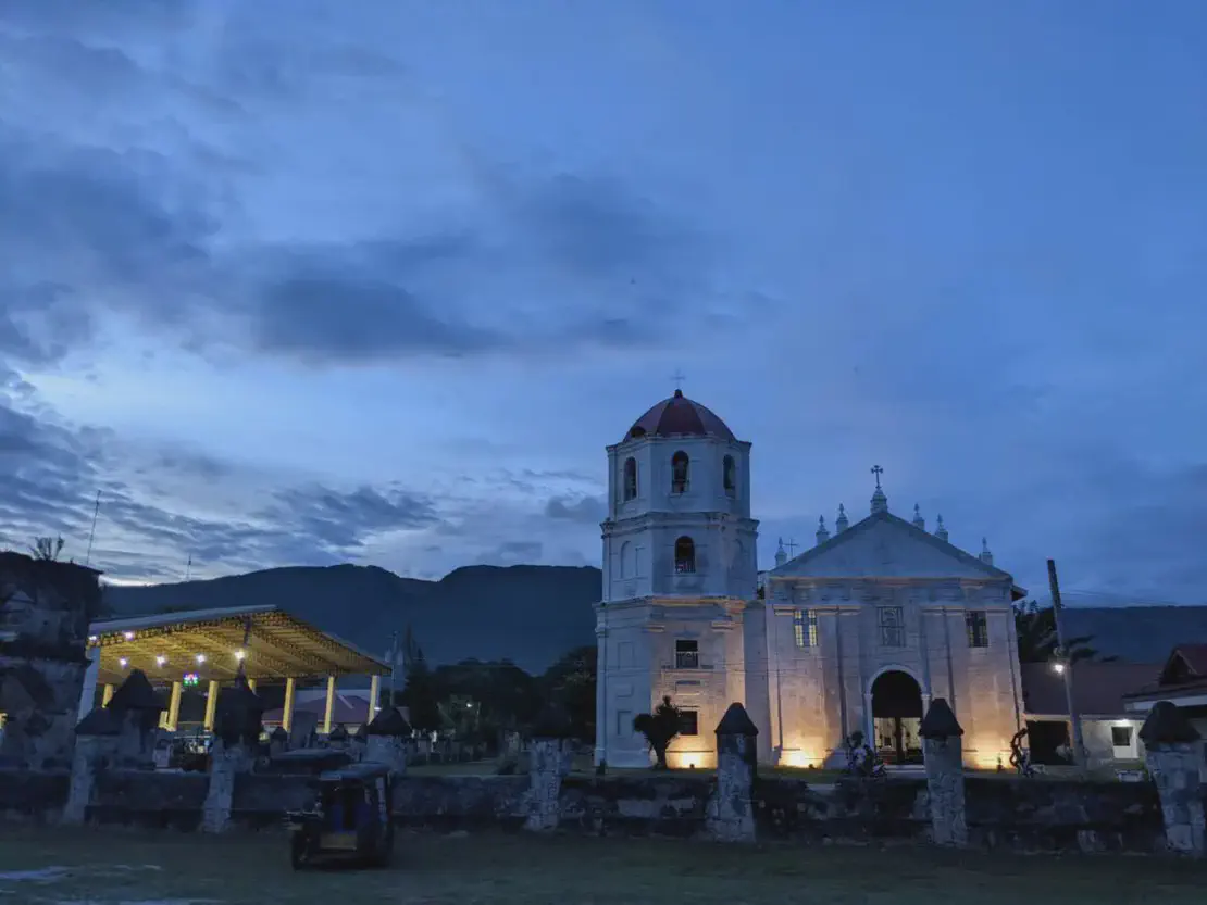 Old stone church with a bell tower illuminated at dusk against a backdrop of mountains.