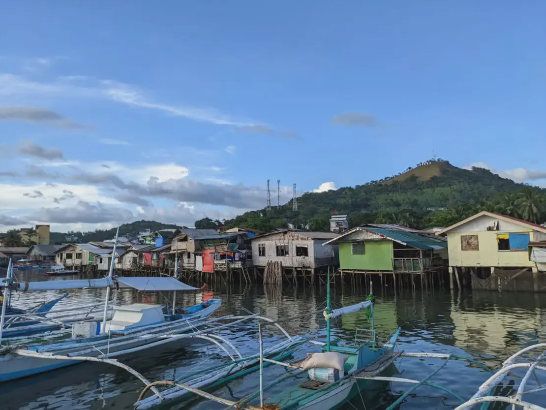 Wooden stilt houses along the water with small fishing boats docked in front and a green hill in the background.