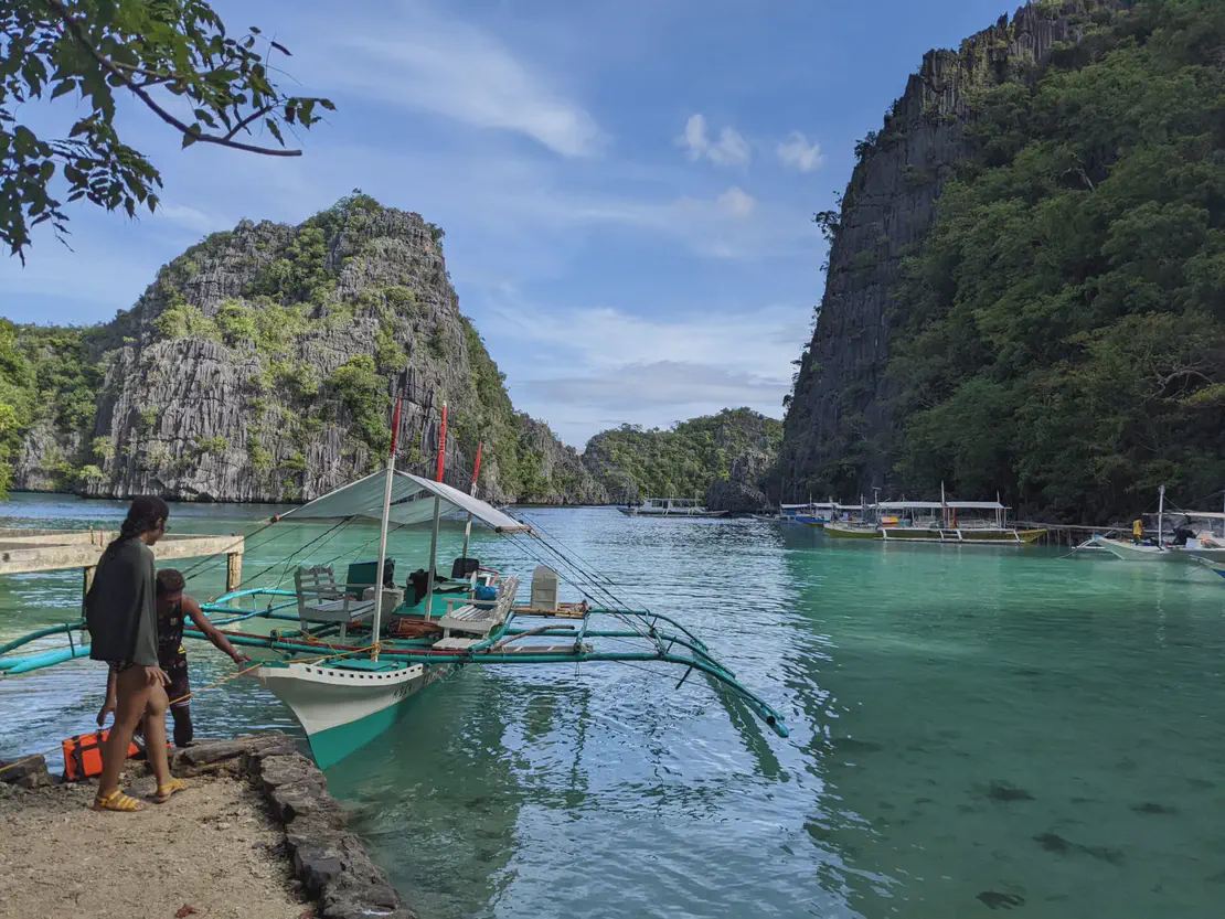 Bangka boat docked on clear turquoise water between limestone cliffs with people standing nearby.