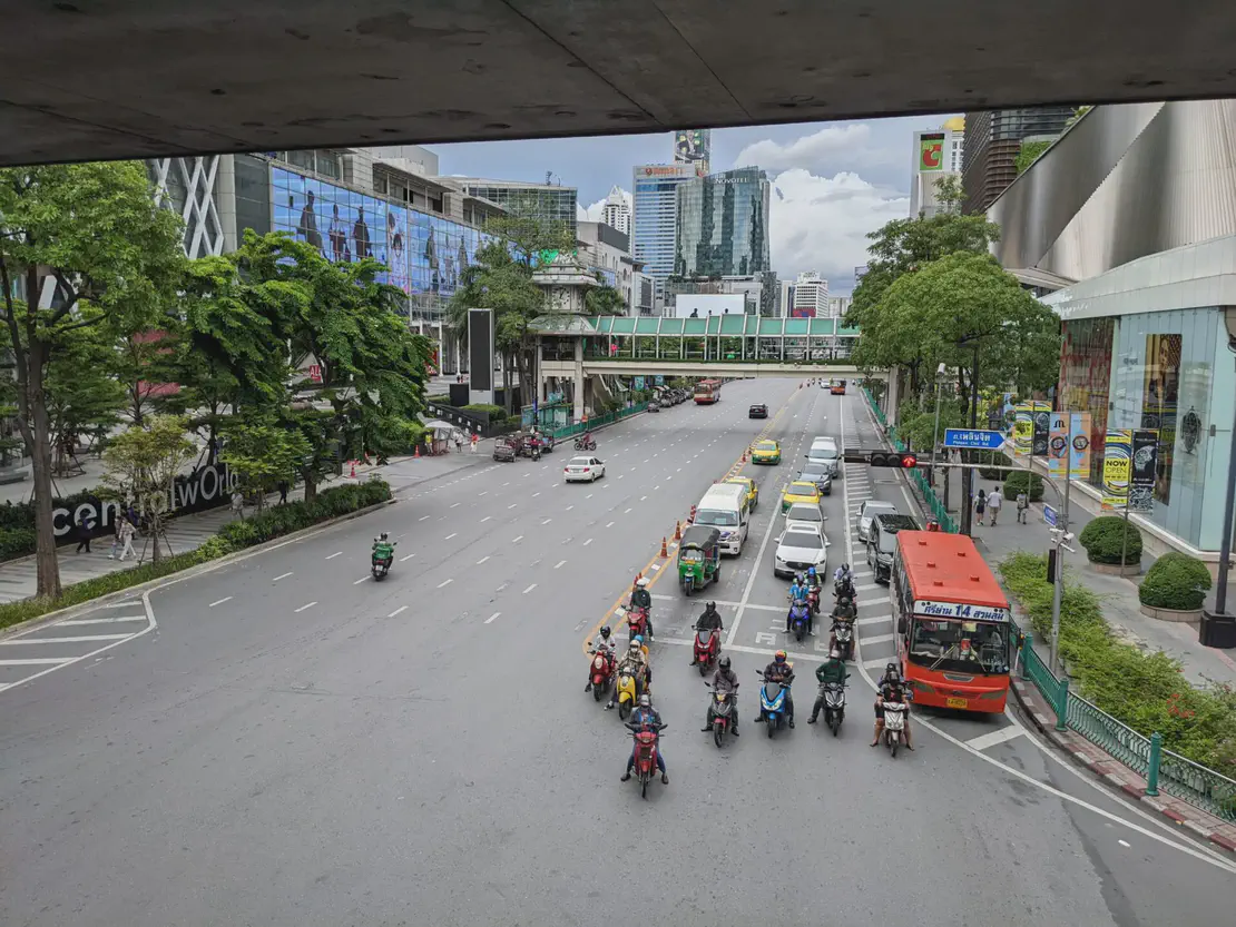 Busy Bangkok street with motorbikes waiting at a traffic light, taxis, a red bus, and shopping malls on both sides.