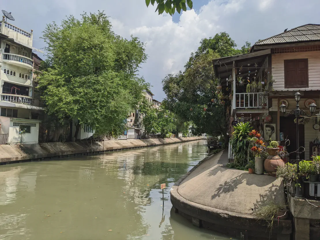 Canal scene with traditional wooden house decorated with plants and portraits, alongside modern buildings and trees in Bangkok.