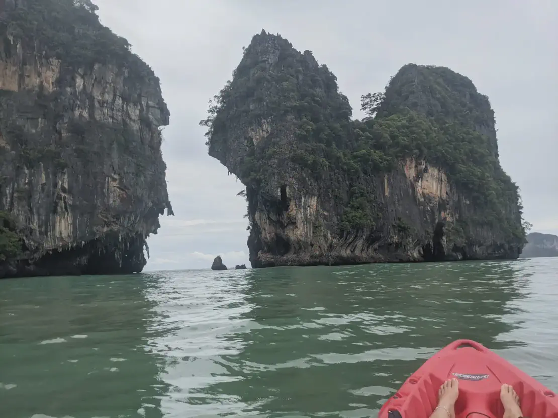 View from a red kayak approaching towering limestone cliffs covered with greenery, with feet visible at the front of the kayak.