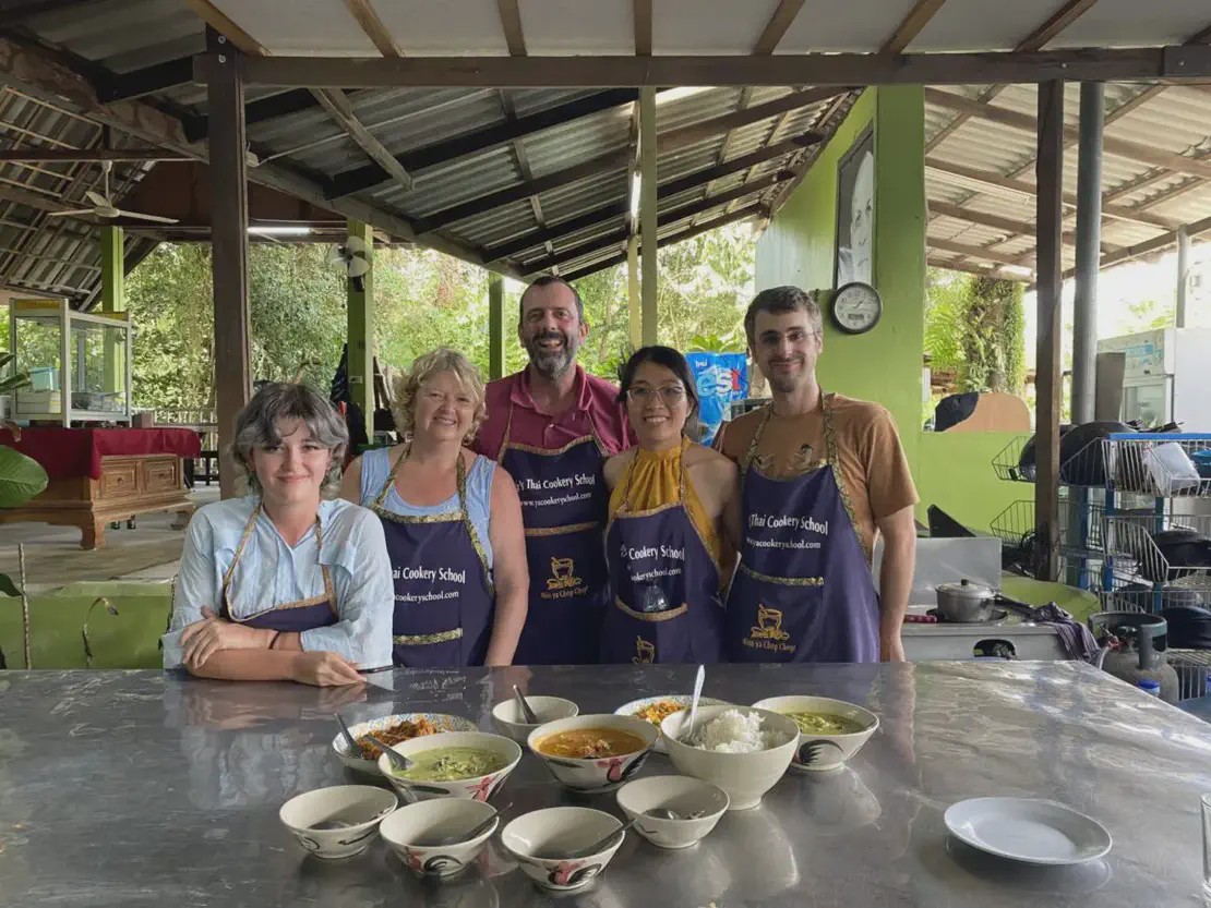 Group of people wearing aprons at a Thai cookery school standing behind a table with bowls of prepared food.
