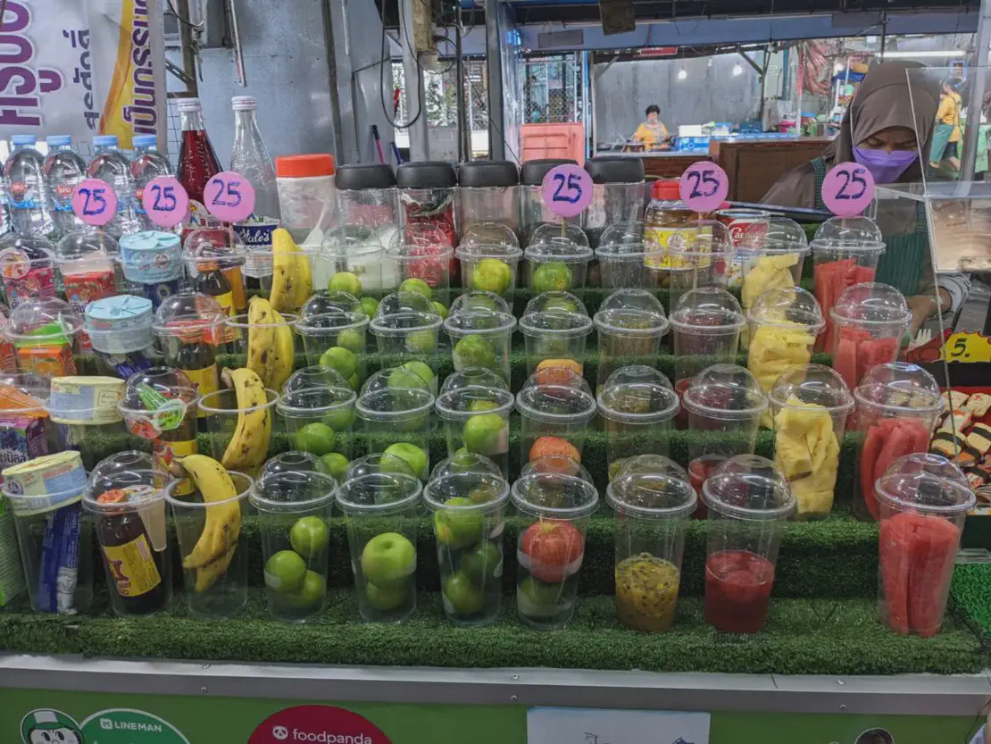 Fruit juice stand with plastic cups filled with fresh fruit such as bananas, apples, watermelon, pineapple, and passionfruit, each priced at 25 baht.