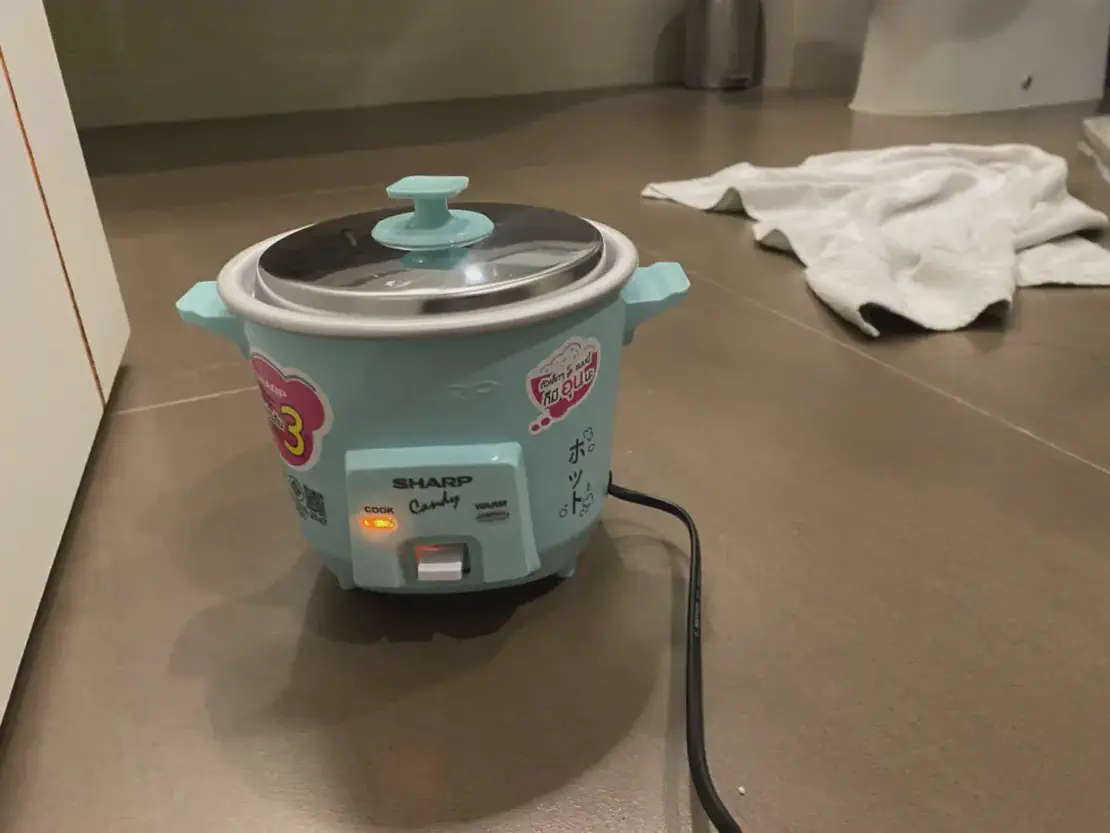 Small turquoise rice cooker with an orange cooking light turned on, placed on a bathroom floor with a towel in the background.