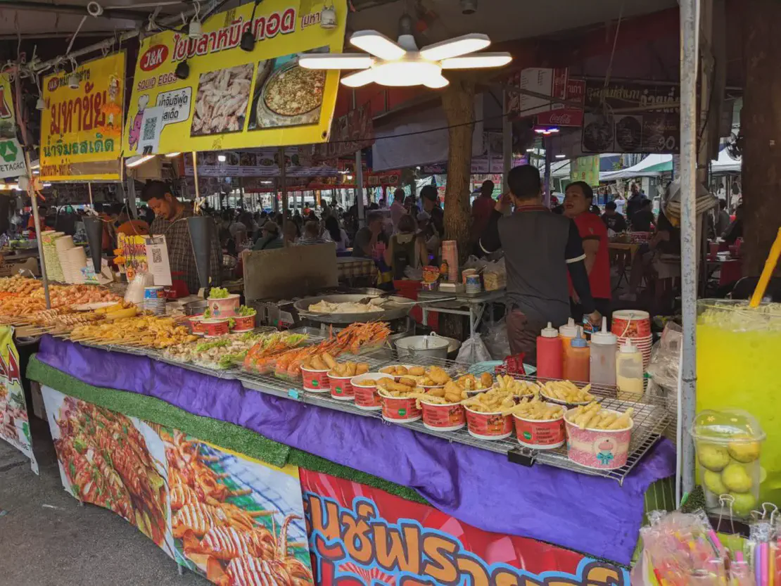 Busy street food market stall with trays of fried snacks, seafood, sauces, and a large container of bright yellow drink.