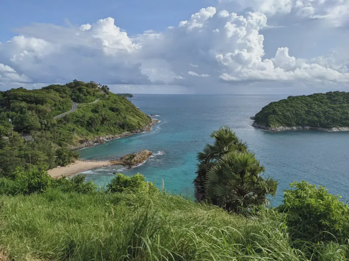 Scenic coastal view of a lush green headland and turquoise sea under a partly cloudy sky, with a small sandy beach below.