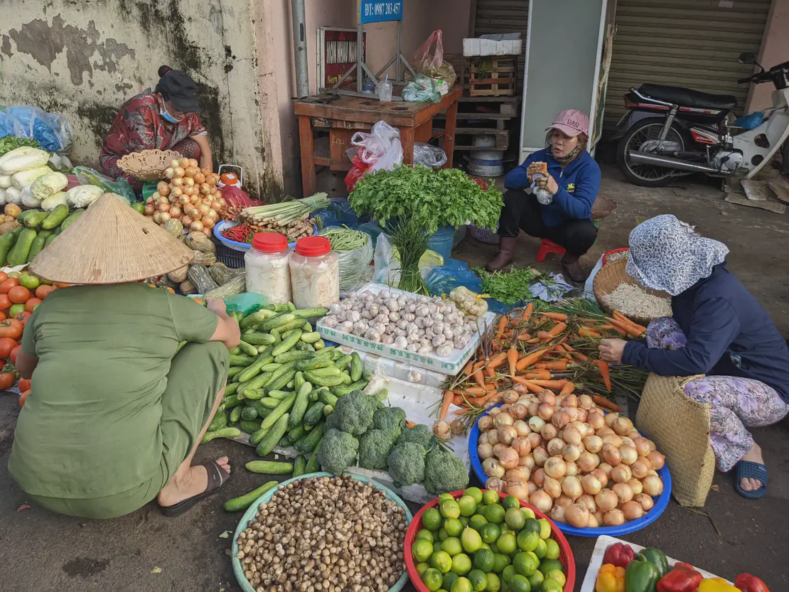 Vendors at an outdoor market selling fresh vegetables including carrots, onions, cucumbers, limes, and broccoli.
