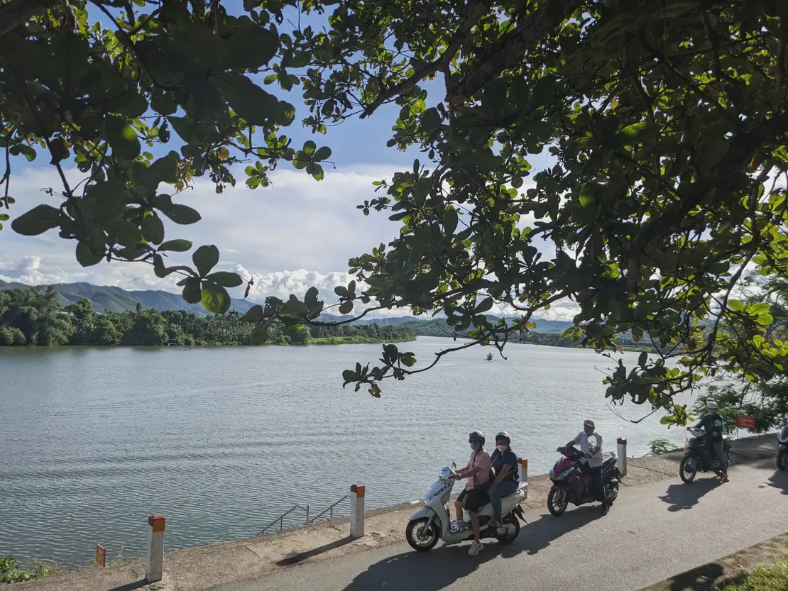 Scooter riders passing along a riverside road with trees framing the view of water and mountains.