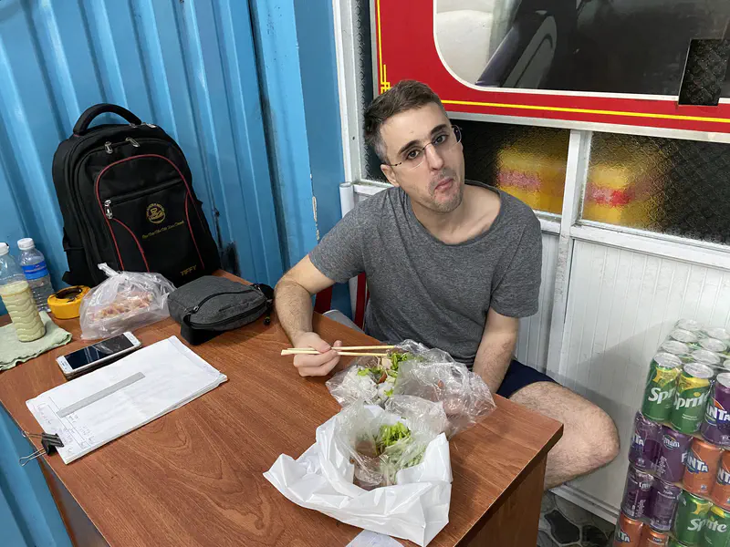 Man eating with chopsticks at a small wooden table inside a shop with snacks and drinks.