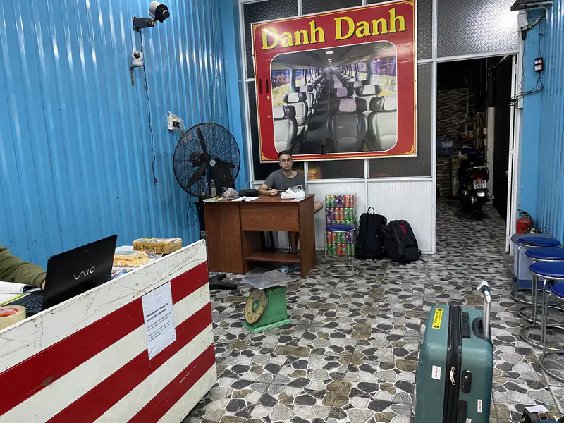 Interior of a bus office with a man eating at a desk, luggage on the floor, and blue walls.