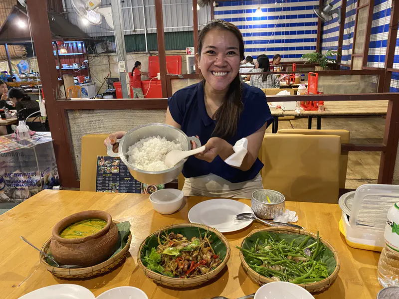 Smiling woman holding a pot of rice at a restaurant table with Cambodian dishes including curry and vegetables.