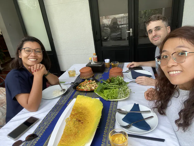 Three friends sitting at a restaurant table with traditional Cambodian dishes including fresh herbs, vegetables, and a large crispy pancake.