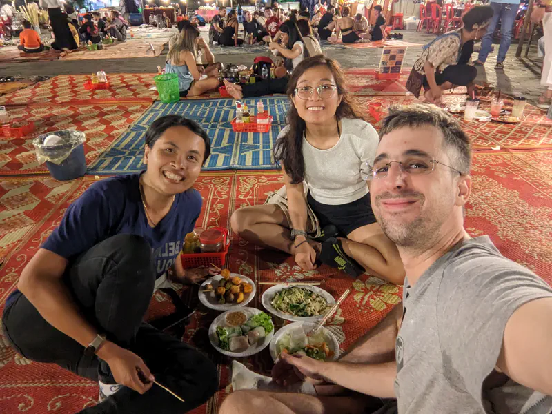 Three friends sitting on mats at a night market, smiling with plates of food in front of them.