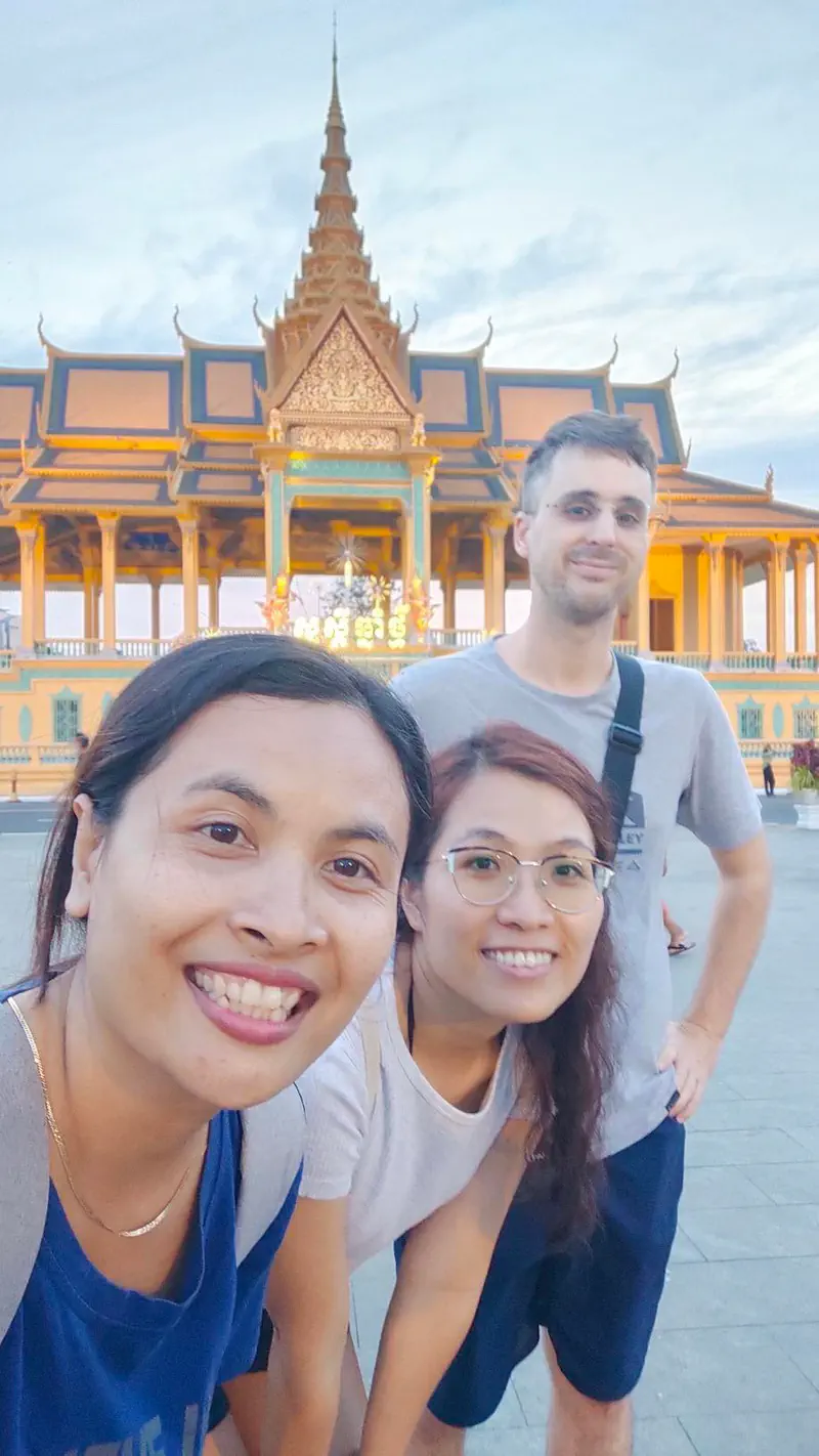 Three friends smiling and posing for a selfie in front of the illuminated Royal Palace in Phnom Penh.