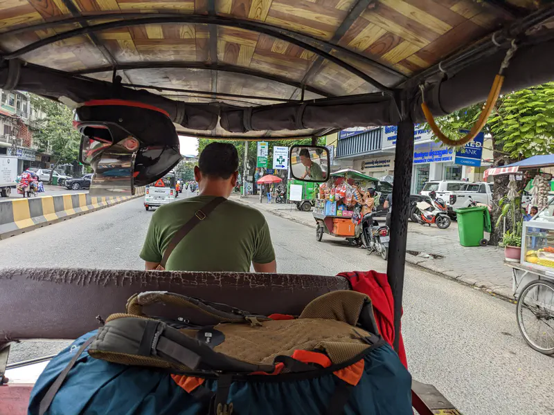 View from the back of a tuk-tuk driving through a busy street with shops, motorbikes, and street vendors.