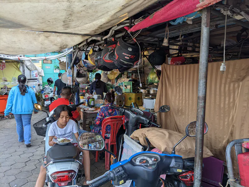 Small food stall with people eating among parked motorbikes under a tarp roof.