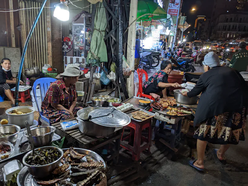 Street food vendors at night with tables full of cooked dishes, pots, and grilled meats, while people sit and prepare food under bright lights.